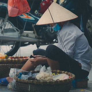 A person wearing a traditional conical hat and a blue face mask is seated on the street, engaging in the task of peeling or preparing food items held in their hands. Surrounding them are baskets filled with various goods, possibly fruits or vegetables. In the background, a motorcycle is partly visible.