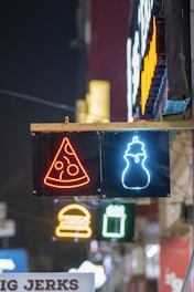 Neon signs hang on a wall, displaying symbols of a pizza slice and a milkshake. Below, other neon signs, such as a burger and a drink, are visible in the background. The scene is illuminated in vivid colors against a dark night setting.