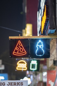 Neon signs hang on a wall, displaying symbols of a pizza slice and a milkshake. Below, other neon signs, such as a burger and a drink, are visible in the background. The scene is illuminated in vivid colors against a dark night setting.