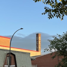 A red brick industrial building is visible with a tall chimney featuring white lettering spelling 'GÉL'. In the foreground, there is a street sign at the intersection of Rachel St and Saint-André, along with some tree branches. The sky is clear with a hint of clouds.