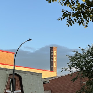 A red brick industrial building is visible with a tall chimney featuring white lettering spelling 'GÉL'. In the foreground, there is a street sign at the intersection of Rachel St and Saint-André, along with some tree branches. The sky is clear with a hint of clouds.