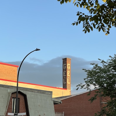 A red brick industrial building is visible with a tall chimney featuring white lettering spelling 'GÉL'. In the foreground, there is a street sign at the intersection of Rachel St and Saint-André, along with some tree branches. The sky is clear with a hint of clouds.