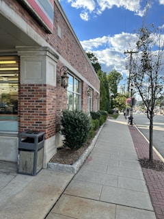 A sidewalk runs alongside a brick building with large windows and an old-fashioned lantern. The environment includes well-maintained bushes and a few young trees. A trash bin is placed near the building's corner. The sky is partly cloudy, with a mix of blue and white clouds.