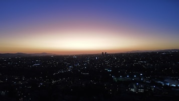 The image captures a panoramic cityscape at dusk, with the skyline outlined against the dimming light of sunset. There is a gradient in the sky transitioning from deep blue at the top to orange and purple near the horizon. The city lights begin to twinkle below, with buildings and streets dimly visible.