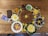 An assortment of foods is arranged on a wooden table. A large round bread decorated with yellow flowers is at the center, surrounded by bowls of various snacks: dried fruits, cheeses, chocolate pieces, and vegetable slices. Colorful plates with cheese slices and flower garnishes add an artistic touch. Chocolate bars and packaging are placed on the side, and small blooms and green leaves accentuate the arrangement.