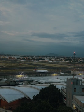 An airport runway with a plane taxiing. The scene includes terminal buildings with arched roofs in the foreground, and a control tower with lights. The background is a cloudy sky with a few distant hills.