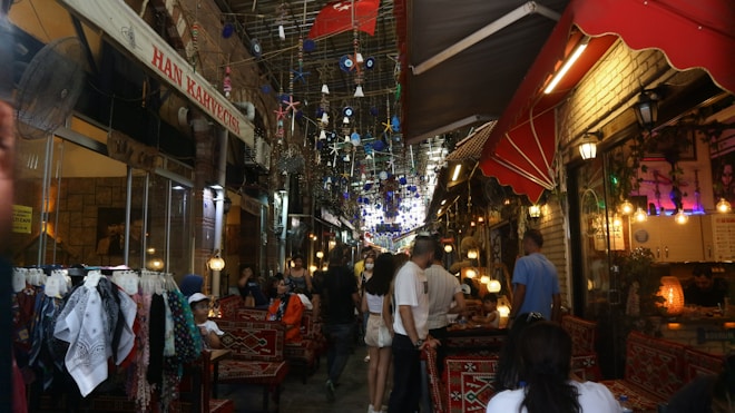 A bustling narrow market street with colorful lanterns and decorations hanging overhead. People are seated and walking around, with shops lining either side selling various textiles and goods. Brightly colored rugs and fabrics are displayed prominently.