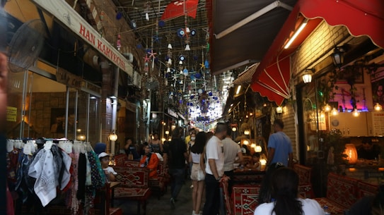 A bustling narrow market street with colorful lanterns and decorations hanging overhead. People are seated and walking around, with shops lining either side selling various textiles and goods. Brightly colored rugs and fabrics are displayed prominently.