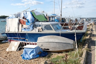 A weathered boat with a blue hull is covered with a variety of tarps and materials, indicating neglect or makeshift repairs. The boat is stationary on a pebble shoreline. Additional boats are visible in the background on the water. The sky is partly cloudy, and there are various items, including an upside-down small boat, scattered nearby.