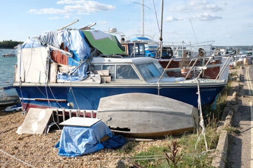 A weathered boat with a blue hull is covered with a variety of tarps and materials, indicating neglect or makeshift repairs. The boat is stationary on a pebble shoreline. Additional boats are visible in the background on the water. The sky is partly cloudy, and there are various items, including an upside-down small boat, scattered nearby.