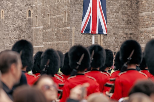 A group of guards wearing traditional red uniforms and black bearskin hats stand in formation. They are facing a large Union Jack flag against the backdrop of a stone wall. The scene appears to be a ceremonial or formal event with onlookers in the foreground.