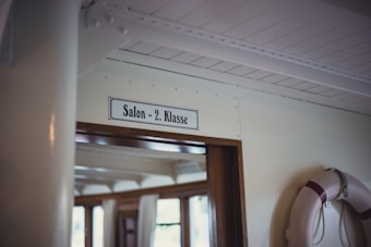 A sign reading 'Salon - 2. Klasse' is mounted above a doorway with wooden trim. The area is inside a ship, indicated by the life preserver hanging on the wall nearby. Soft natural light enters through windows, highlighting the interior maritime decor.