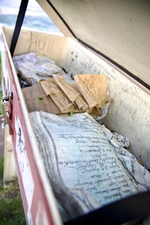 A weathered wooden box contains various aged papers and scrolls with handwritten notes. The interior walls of the box are inscribed with additional writing. The box is open, revealing its contents, which appear historical or personal in nature. The scene conveys a sense of curiosity and nostalgia.