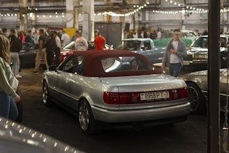 An indoor parking area filled with several vintage and classic cars. A silver convertible with a red roof is prominently displayed in the foreground. The area is dimly lit with string lights hanging from the ceiling, and a crowd of people, casually dressed, are gathered around, possibly attending a car show.