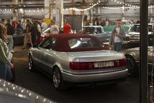 An indoor parking area filled with several vintage and classic cars. A silver convertible with a red roof is prominently displayed in the foreground. The area is dimly lit with string lights hanging from the ceiling, and a crowd of people, casually dressed, are gathered around, possibly attending a car show.