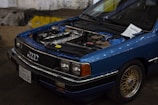 A blue Audi car with its hood open, revealing the engine. The front grille displays the Audi logo, and a note is placed on the windshield. The car has a California license plate and is parked in a dimly lit indoor area with a rough concrete floor.
