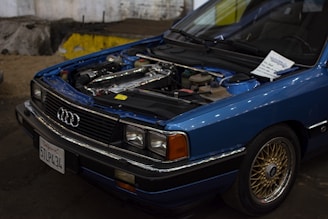 A blue Audi car with its hood open, revealing the engine. The front grille displays the Audi logo, and a note is placed on the windshield. The car has a California license plate and is parked in a dimly lit indoor area with a rough concrete floor.