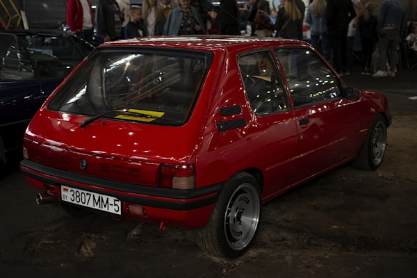 A red vintage car is parked indoors on a slightly cracked concrete surface. The car's rear and side are prominently visible, showcasing the Peugeot 205 model identification and a license plate from Belarus. Surrounding the car, some people are standing, suggesting an exhibition or gathering.