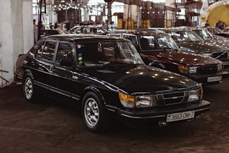 Several classic cars are parked closely together inside a large industrial-style building. The focus is on a black Saab Turbo sedan with vintage features and a Belarusian license plate. The surroundings are dimly lit with artificial lighting reflecting off the cars' surfaces. Other cars of similar vintage style are visible in the background, adding a nostalgic atmosphere to the scene.