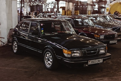 Several classic cars are parked closely together inside a large industrial-style building. The focus is on a black Saab Turbo sedan with vintage features and a Belarusian license plate. The surroundings are dimly lit with artificial lighting reflecting off the cars' surfaces. Other cars of similar vintage style are visible in the background, adding a nostalgic atmosphere to the scene.