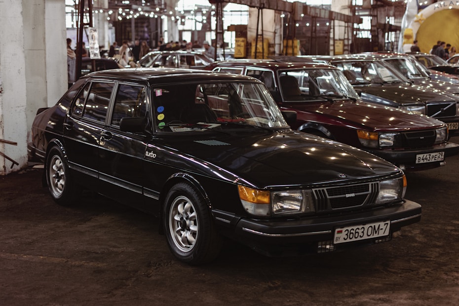 Several classic cars are parked closely together inside a large industrial-style building. The focus is on a black Saab Turbo sedan with vintage features and a Belarusian license plate. The surroundings are dimly lit with artificial lighting reflecting off the cars' surfaces. Other cars of similar vintage style are visible in the background, adding a nostalgic atmosphere to the scene.