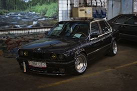 A classic black BMW sedan is parked indoors, showcasing a sleek, polished exterior with custom rims and yellow-tinted headlights. The license plate is visible, and there is a painted backdrop of a river and forest scene on the wall behind the car. The environment resembles a garage or auto shop, with various equipment and industrial elements in the background.