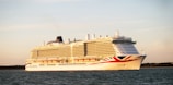 A large cruise ship is sailing on a calm body of water during sunset. The ship is white with red and blue accents and has multiple decks with rows of windows. The sky is clear with a warm, golden hue.