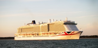 A large cruise ship is sailing on a calm body of water during sunset. The ship is white with red and blue accents and has multiple decks with rows of windows. The sky is clear with a warm, golden hue.