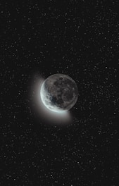 A celestial scene features the moon partially illuminated against a vast, starry night sky. The surface of the moon reveals craters and a subtle glow emanates from its lit side, creating a serene and captivating contrast with the surrounding darkness.