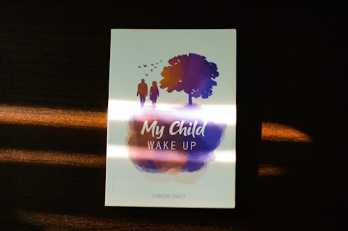 A book cover resting on a wooden surface, featuring the title 'My Child Wake Up.' The design includes silhouettes of a man and a child walking towards a tree with birds flying overhead, all depicted in purple and orange hues.