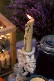 A lit candle with melted wax drips is placed in an intricately designed holder resembling a mythical creature. Surrounding the candle is a setting with elements like a bundle of corks in a wooden frame, a glass bottle with twine around its neck, and a jar with a metal clasp lid. In the background, a bouquet of purple flowers adds a pop of color.