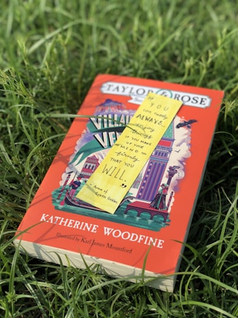 A book titled 'Katherine Woodfine' is lying in a grassy field. The cover features colorful illustrations with a dominant red background, showcasing a variety of architectural elements. A yellow sticky note with a quote from 'Anne of Green Gables' is placed on top of the book.