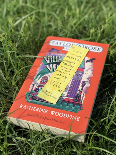 A book titled 'Katherine Woodfine' is lying in a grassy field. The cover features colorful illustrations with a dominant red background, showcasing a variety of architectural elements. A yellow sticky note with a quote from 'Anne of Green Gables' is placed on top of the book.