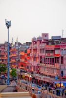 A vibrant market street in Jaipur, India, featuring traditional Rajasthani architecture with pink and red buildings illuminated by warm lighting. The street is bustling with people, vehicles, and shops creating an energetic urban atmosphere.