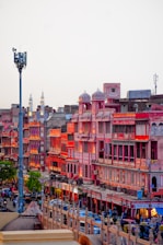 A vibrant market street in Jaipur, India, featuring traditional Rajasthani architecture with pink and red buildings illuminated by warm lighting. The street is bustling with people, vehicles, and shops creating an energetic urban atmosphere.
