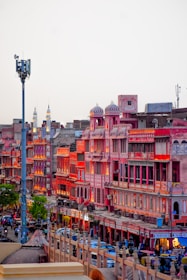 A vibrant market street in Jaipur, India, featuring traditional Rajasthani architecture with pink and red buildings illuminated by warm lighting. The street is bustling with people, vehicles, and shops creating an energetic urban atmosphere.