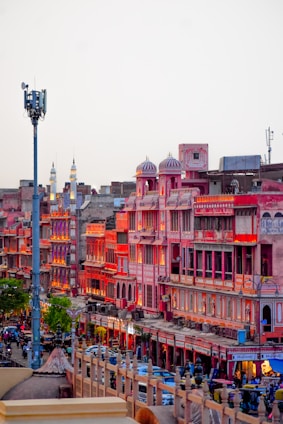 A vibrant market street in Jaipur, India, featuring traditional Rajasthani architecture with pink and red buildings illuminated by warm lighting. The street is bustling with people, vehicles, and shops creating an energetic urban atmosphere.