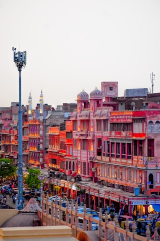 A vibrant market street in Jaipur, India, featuring traditional Rajasthani architecture with pink and red buildings illuminated by warm lighting. The street is bustling with people, vehicles, and shops creating an energetic urban atmosphere.