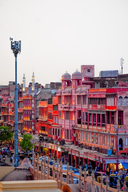 A vibrant market street in Jaipur, India, featuring traditional Rajasthani architecture with pink and red buildings illuminated by warm lighting. The street is bustling with people, vehicles, and shops creating an energetic urban atmosphere.