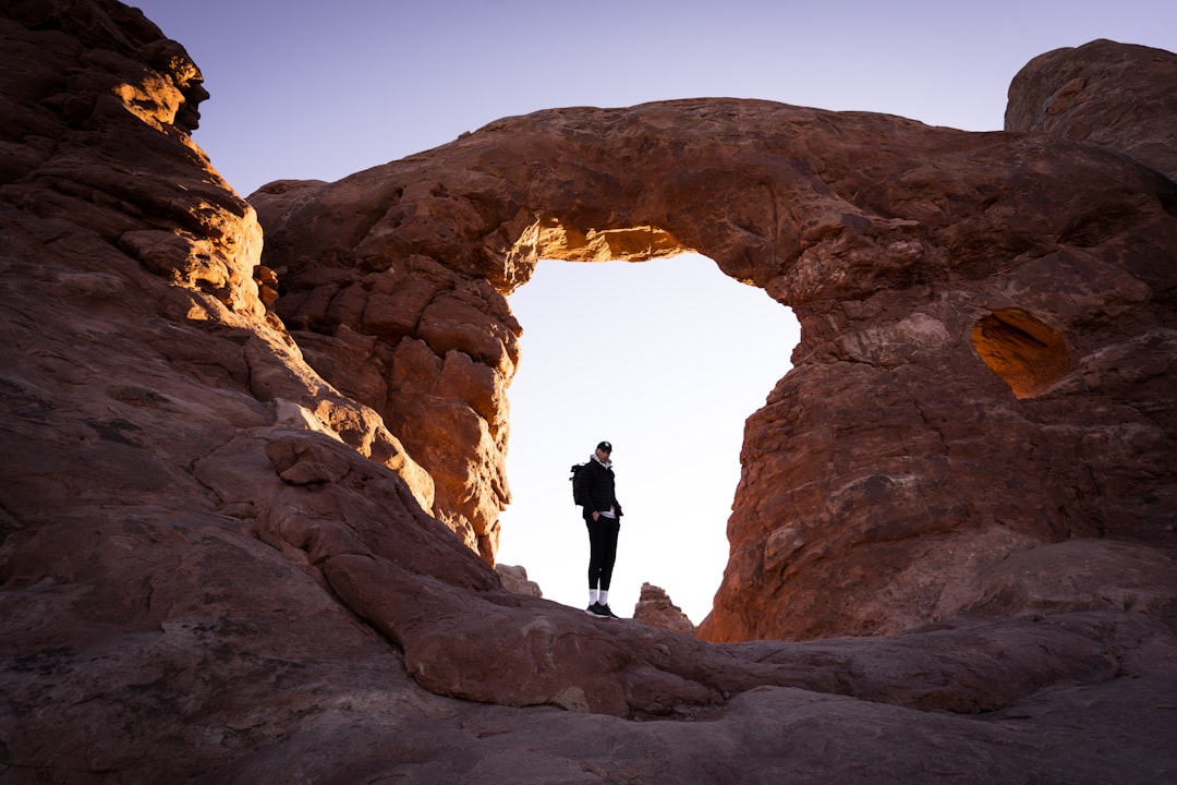 a person standing in a large rock cave, A lone hiker looks back at the camera in Arches National Park at sunrise