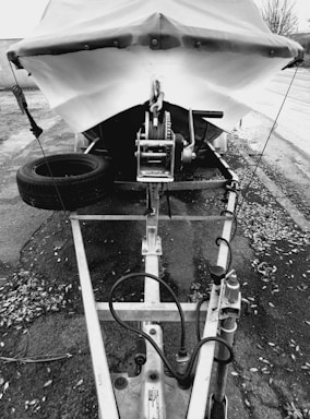 A black and white image of a covered boat on a trailer, with a tire attached to the side. The trailer is positioned on a paved surface surrounded by fallen leaves. The boat is secured by a winch, with visible ropes and pulleys.