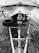 A black and white image of a covered boat on a trailer, with a tire attached to the side. The trailer is positioned on a paved surface surrounded by fallen leaves. The boat is secured by a winch, with visible ropes and pulleys.