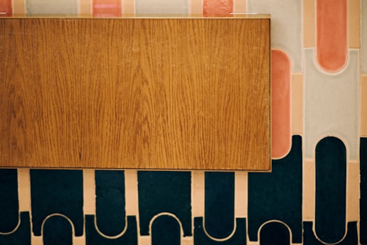 A wooden rectangular panel is placed against a decorative wall featuring an abstract pattern with rounded shapes and strips in shades of pink, white, and dark teal.