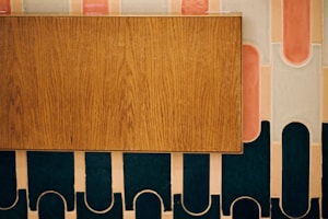 A wooden rectangular panel is placed against a decorative wall featuring an abstract pattern with rounded shapes and strips in shades of pink, white, and dark teal.