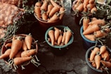 Multiple buckets filled with fresh, orange carrots are arranged on a concrete surface. Some carrots still have green tops attached. A mesh bag containing additional carrots is partially visible in the background.