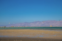 A tranquil beach scene with clear blue skies and vibrant turquoise waters. Several colorful kites are seen above the water, suggesting kite surfing or similar activities. A range of mountains is visible in the distance, providing a stunning backdrop. The sandy beach in the foreground is expansive and mostly flat, indicating low tide.