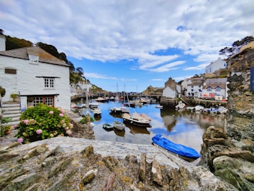 A picturesque coastal village with a serene harbor filled with small boats. Charming white cottages with thatched roofs line the waterfront, surrounded by rocky cliffs and lush greenery. The sky is partly cloudy, providing a vibrant blue backdrop reflected in the calm water.