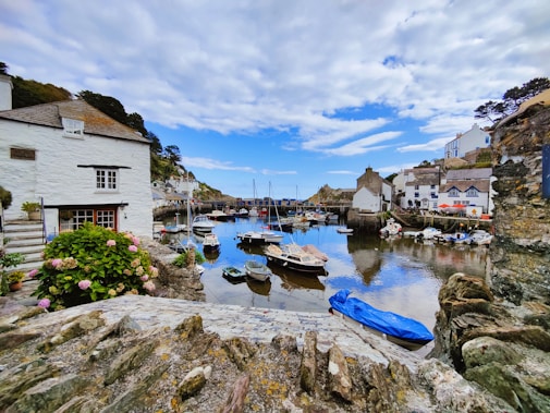 A picturesque coastal village with a serene harbor filled with small boats. Charming white cottages with thatched roofs line the waterfront, surrounded by rocky cliffs and lush greenery. The sky is partly cloudy, providing a vibrant blue backdrop reflected in the calm water.