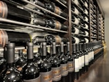 A well-organized wine cellar with rows of wine bottles stored horizontally on wooden racks. The bottles have elegant labels, indicating different varieties and vintages. The setting is dimly lit, suggesting a sophisticated, serene atmosphere typically associated with wine storage.