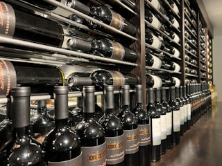 A well-organized wine cellar with rows of wine bottles stored horizontally on wooden racks. The bottles have elegant labels, indicating different varieties and vintages. The setting is dimly lit, suggesting a sophisticated, serene atmosphere typically associated with wine storage.